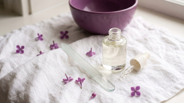 Lima di vetro e olio per cuticole su lino chiaro con petali lilla